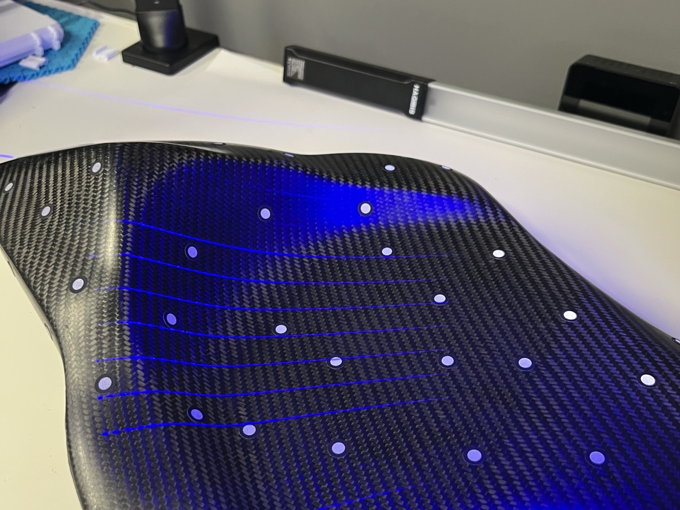 A carbon-fibre motorcycle seat on the scanner bed, marked with tracking dots under structured-light scan lines